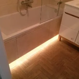 Nowoczesna łazienka we Wrocławiu z podświetlaną wanną obudowaną beżowymi płytkami i drewnopodobną podłogą ułożoną w jodełkę, obok biała szafka umywalkowa.