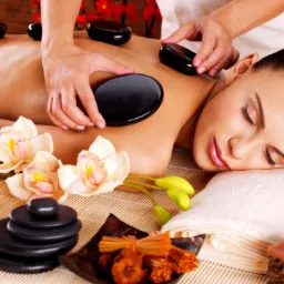 Kobieta leżąca na stole do zabiegów, poddawana terapii gorącymi kamieniami; widoczne czarne, owalne kamienie na plecach, kwiaty orchidei, palące się świece i stos kamieni obok, dłonie terapeuty...