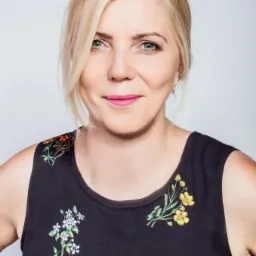 Portret uśmiechniętej blondynki w czarnej sukience w kwiaty na jasnym tle.