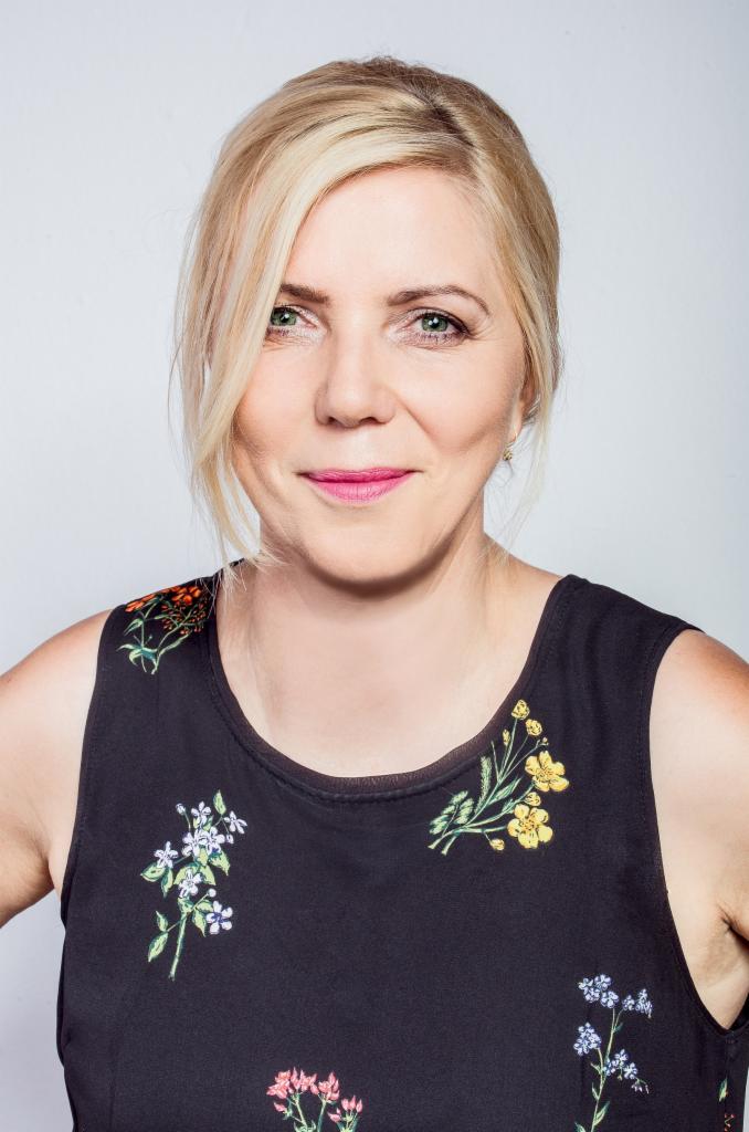 Portret uśmiechniętej blondynki w ciemnej sukience w kwiaty na jasnym tle.