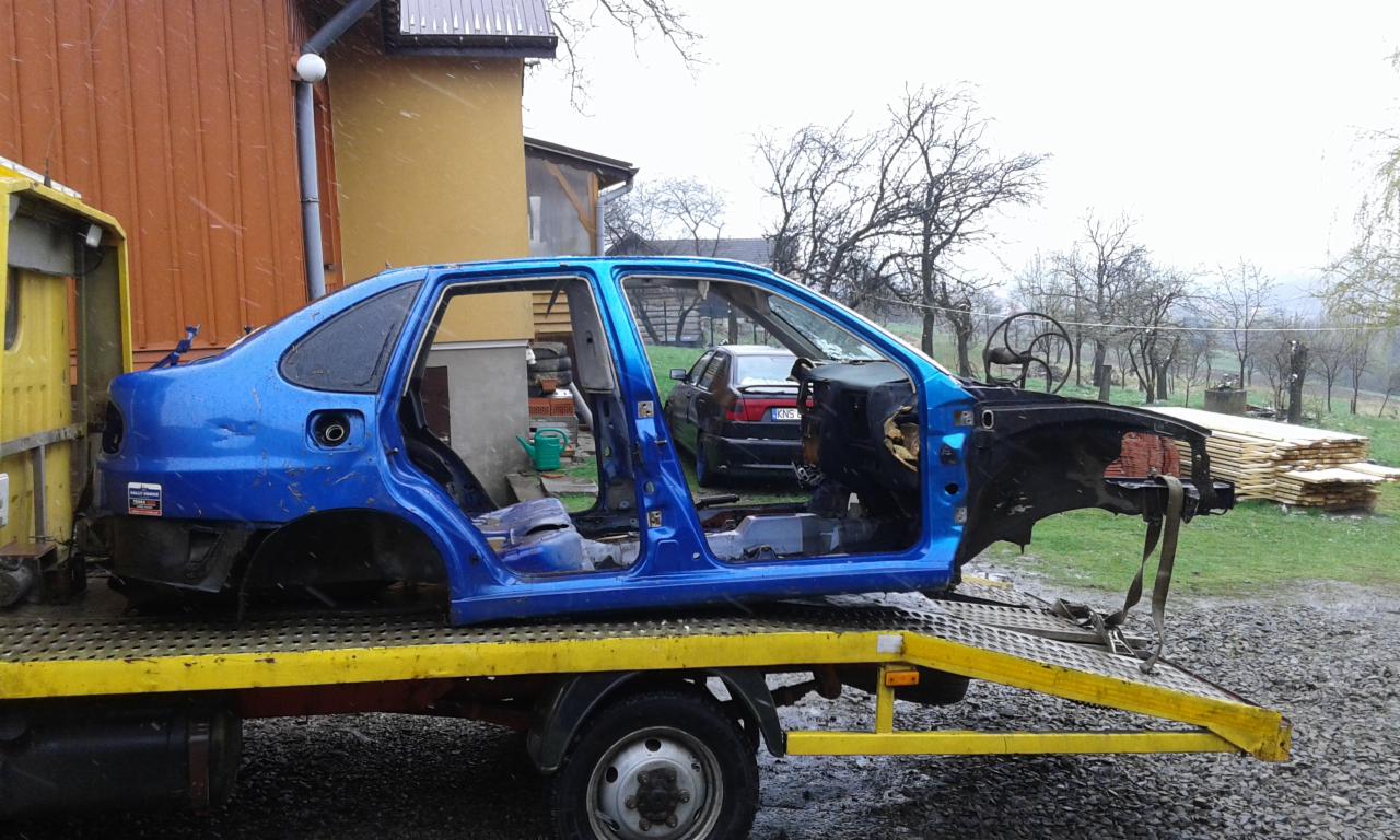 Uszkodzony, niebieski szkielet samochodu bez drzwi i przedniej części, przewożony na żółtej platformie transportowej. W tle widoczny budynek i drzewa.