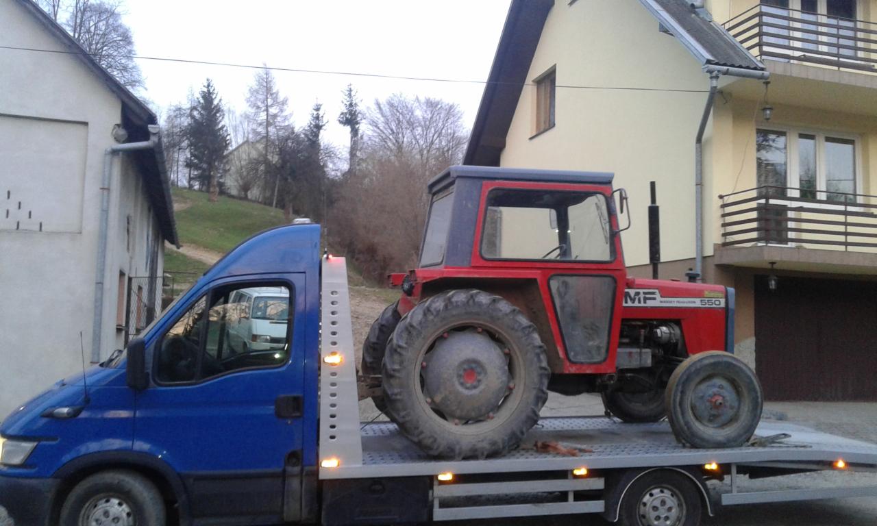 Niebieska laweta przewozi czerwony traktor Massey Ferguson 550 na tle zabudowań.