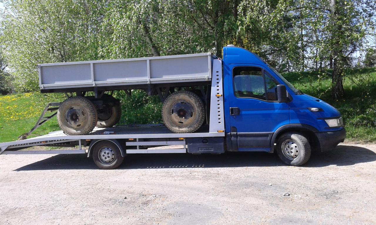 Niebieska ciężarówka z platformą, na której przewożona jest szara przyczepa rolnicza z brudnymi kołami, na tle zieleni.