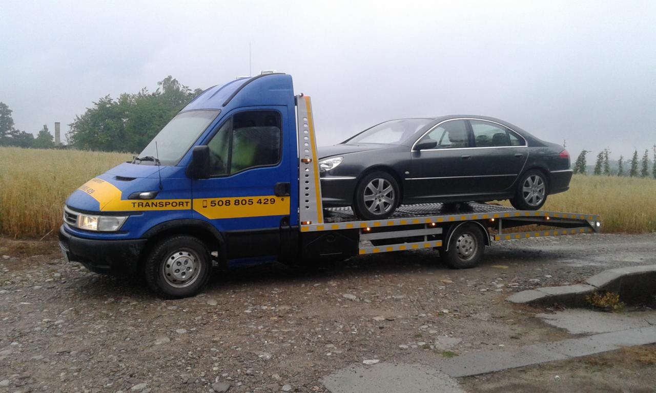 Niebiesko-żółty pojazd transportowy z szarym samochodem osobowym na platformie, widoczne numery telefonu, w tle pole zboża i pochmurne niebo.