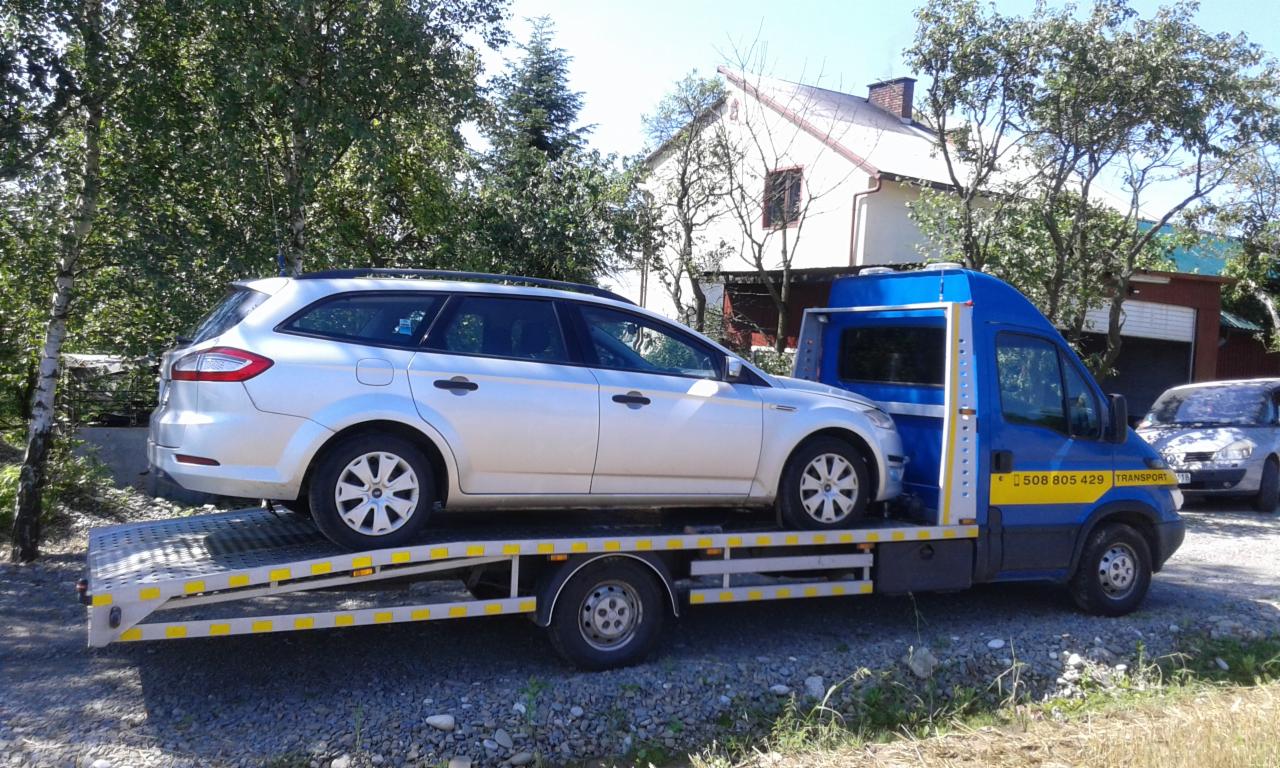 Srebrne kombi Ford Mondeo na platformie niebieskiej ciężarówki pomocy drogowej z żółtymi akcentami, widok z boku na tle drzew i domu.