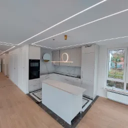 Nowoczesna, jasna kuchnia z wyspą, oświetleniem ledowym w suficie i marmurowym blatem, widok na ogród przez duże okno.