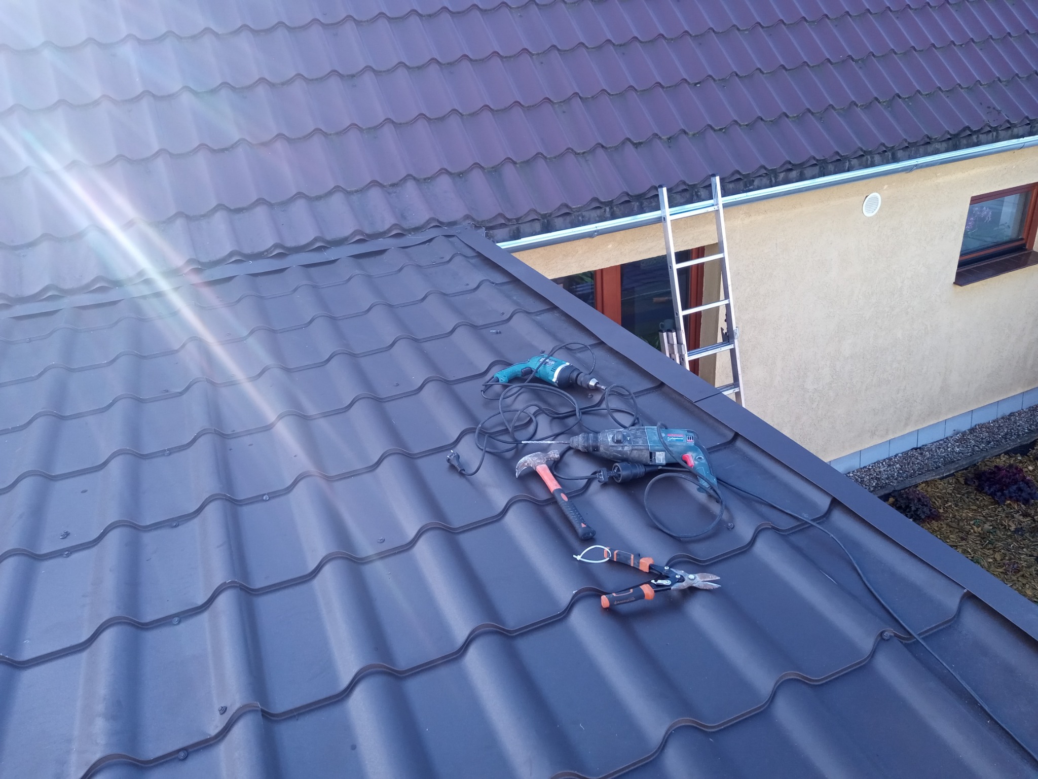 Montaż ciemnej blachodachówki: wiertarka, młotek i nożyce dekarskie na połaci dachu w promieniach słońca, drabina oparta o budynek w tle.