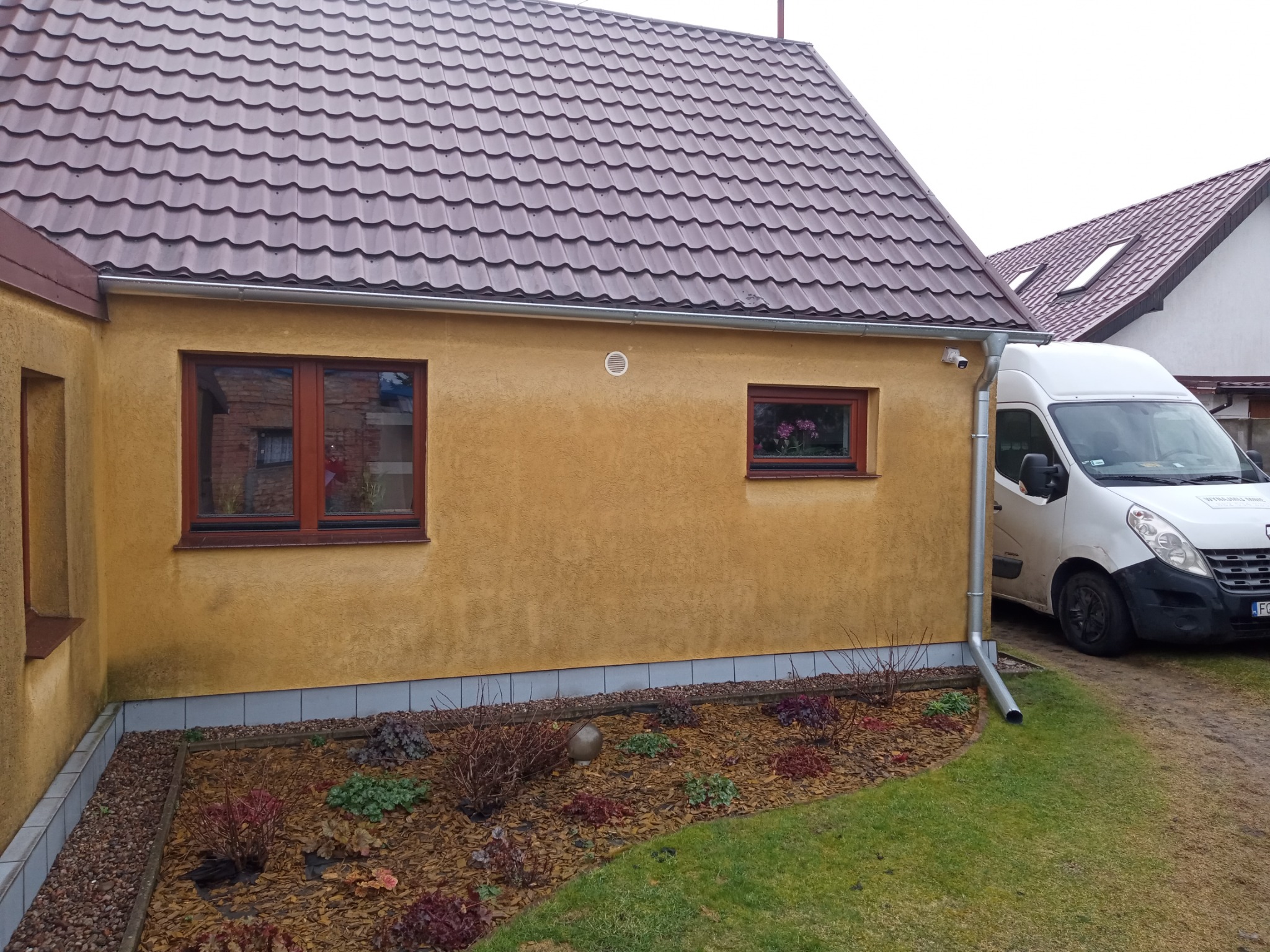 Elewacja domu o żółtej barwie z brązowymi oknami, brązowym dachem i srebrną rynną, obok zaparkowany biały samochód dostawczy.