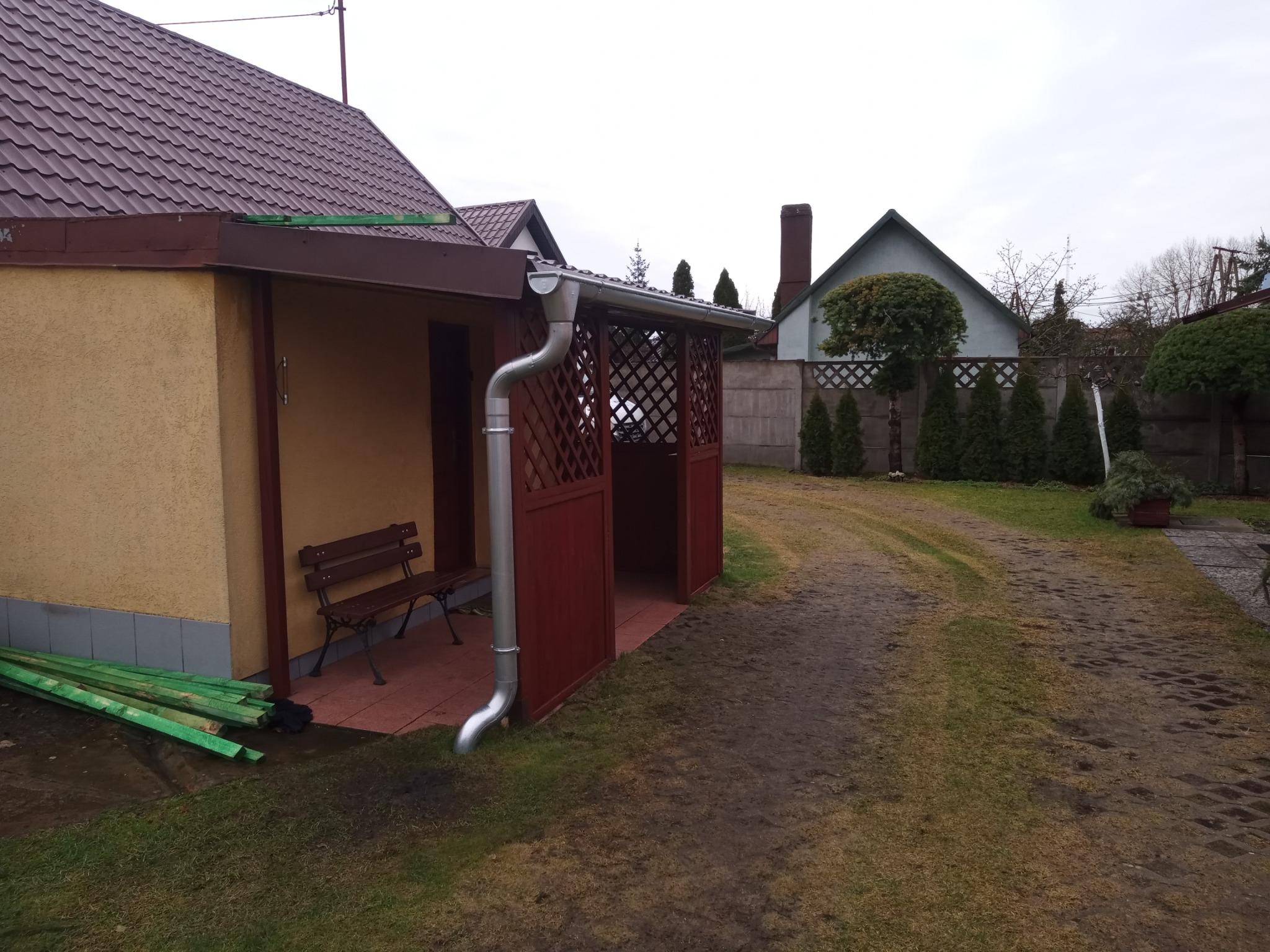 Zewnętrzny widok na budynek z dobudowaną altaną z kratownicą, nowa rynna spustowa, obok ławka i stos zielonych desek, na podłodze terakota.
