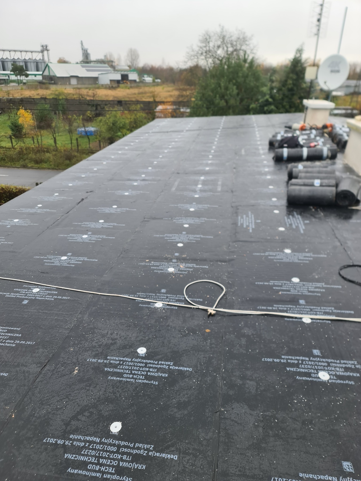 Czarny dach pokryty papą z widocznymi oznaczeniami producenta i białymi punktami mocującymi, z rolkami papy w tle i widokiem na okoliczne budynki i drzewa w oddali.