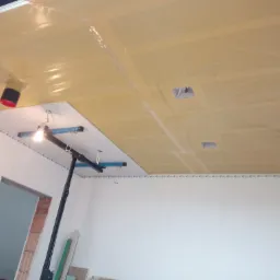 Montaż sufitu podwieszanego: widok na częściowo zamontowany sufit z folią ochronną, widoczne otwory na oświetlenie i narzędzie do montażu sufitów.