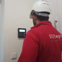 Instalator w kasku konfiguruje panel alarmowy na ścianie, widoczny fragment czerwonej bluzy z logo firmy.