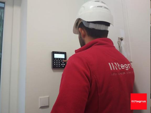 Instalator w kasku konfiguruje panel alarmowy na ścianie, widoczny fragment czerwonej bluzy z logo firmy.