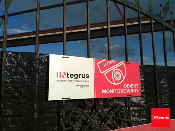 Tablica informacyjna z logotypem firmy Integrus i napisem 'Obiekt Monitorowany' umieszczona na metalowej bramie z widoczną siatką maskującą w tle.