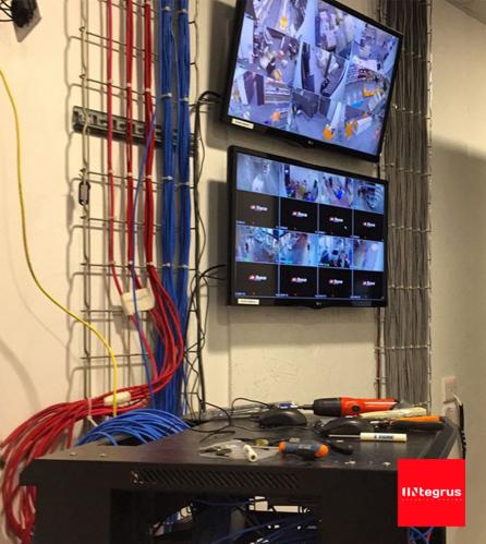 Dwa monitory CCTV zawieszone na ścianie, wyświetlające obraz z kamer. Obok, metalowa drabinka kablowa z poprowadzonymi kablami w kolorach czerwonym i niebieskim. Na szafce RACK leżą narzędzia...
