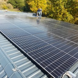 4EL - Instalacja paneli fotowoltaicznych na blaszanym dachu, monter w oddali zabezpiecza połączenia, jesienne drzewa w tle.