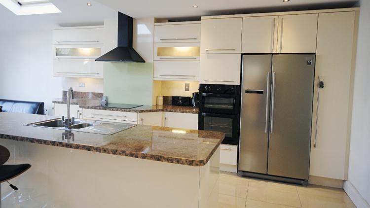 Jasna kuchnia z beżowymi szafkami, czarnym okapem i blatem z brązowego kamienia, widok na dwudrzwiową lodówkę ze stali nierdzewnej.