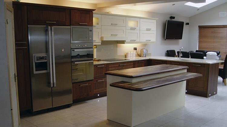 Nowoczesna kuchnia z dwupoziomową wyspą, zabudową w odcieniach brązu i bieli, oraz sprzętem AGD ze stali nierdzewnej.