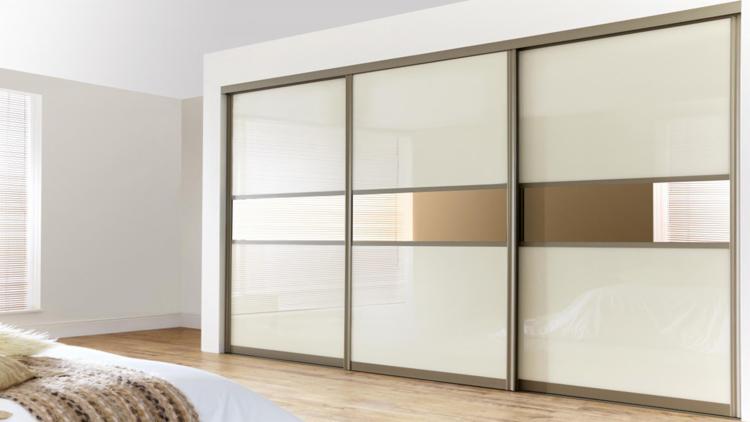 Nowoczesna szafa przesuwna z beżowymi panelami i poziomymi lustrzanymi wstawkami, widoczna w jasnym wnętrzu sypialni z fragmentem łóżka w dolnym rogu.