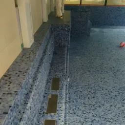 Prace wykończeniowe przy basenie z mozaiką w odcieniach błękitu, widoczna osoba w kombinezonie ochronnym i piłka do zabawy.