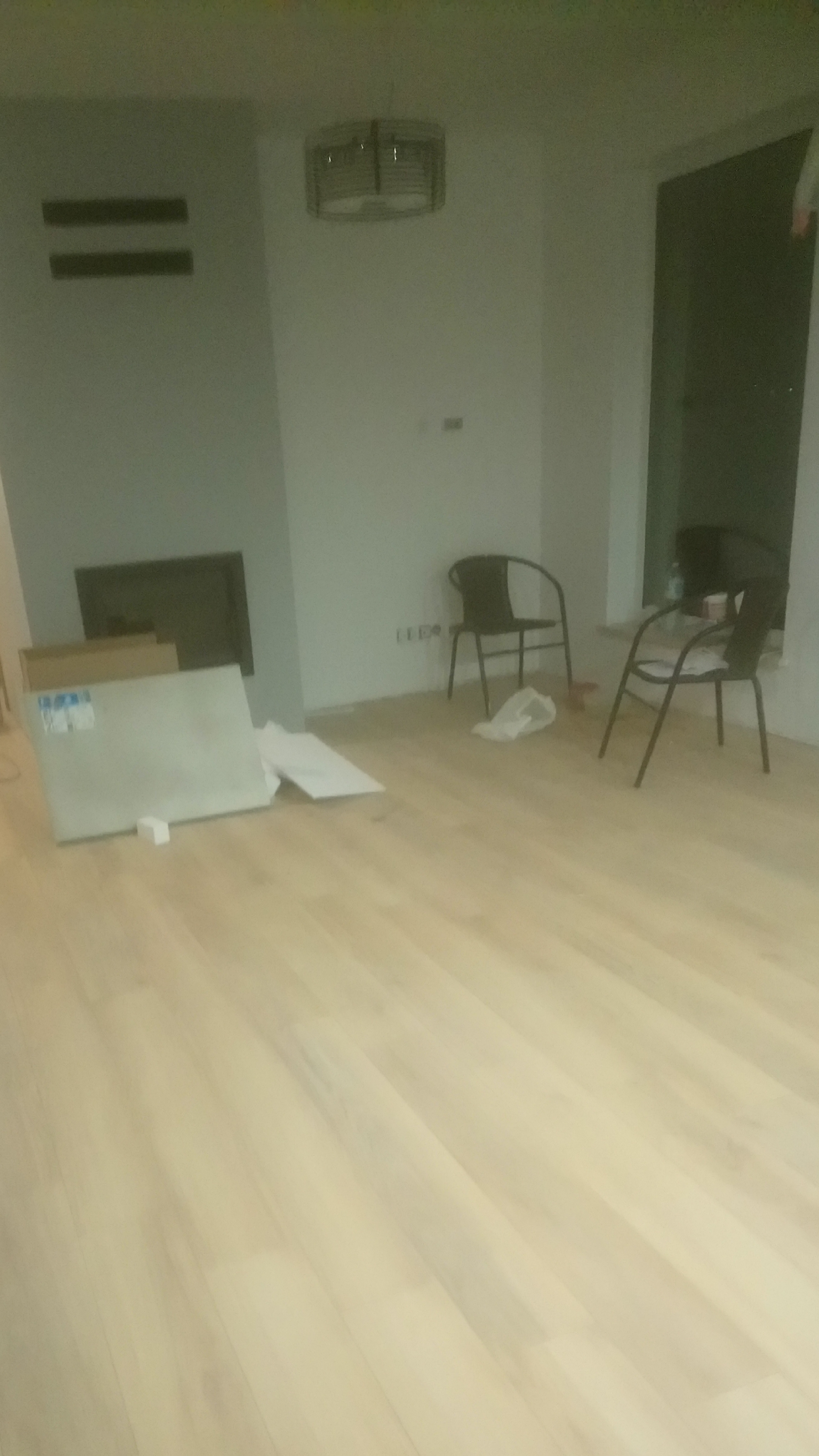 Jasne panele podłogowe w trakcie układania, widoczne paczki z panelami, dwa krzesła i minimalistyczna lampa sufitowa w tle