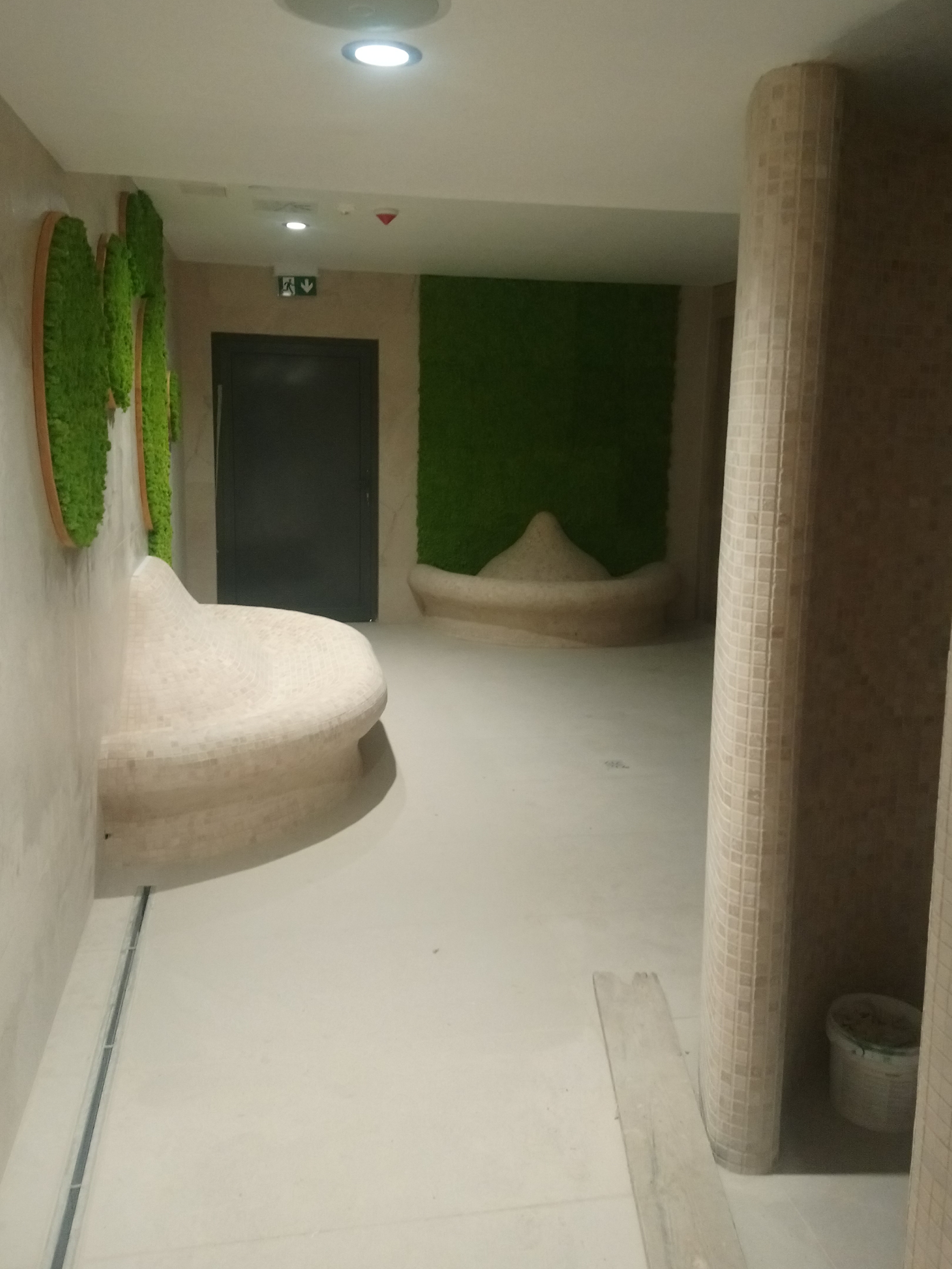 Wnętrze spa w Ostrowie Wielkopolskim z mozaikową ławką i kolumnami, zieloną ścianą z mchu i minimalistycznym oświetleniem sufitowym.