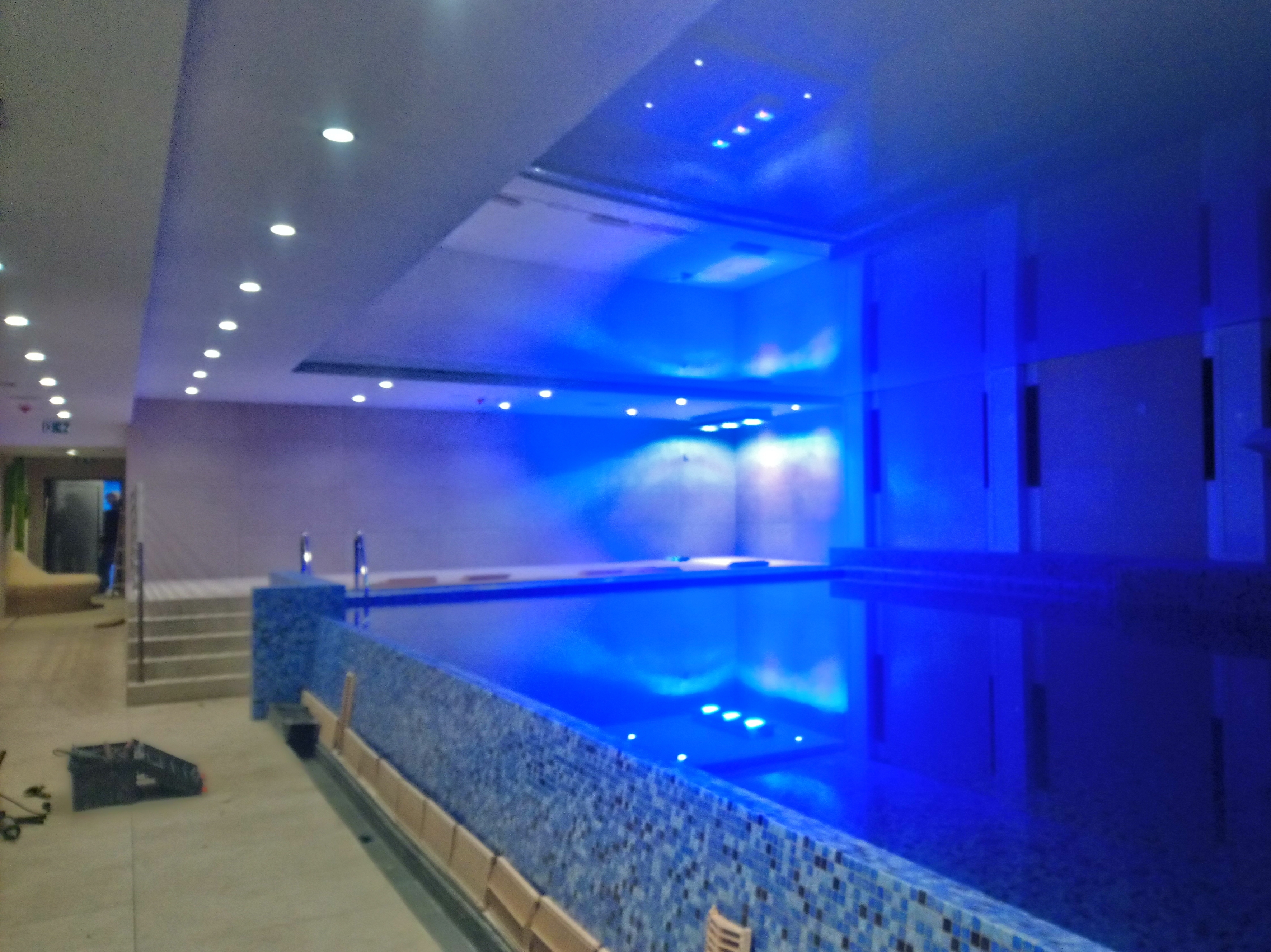 Nowoczesny basen kryty z mozaiką w odcieniach błękitu, oświetlony punktowymi lampami sufitowymi i niebieskim światłem, z widocznymi schodami wejściowymi i elementami wykończeniowymi w trakcie prac.