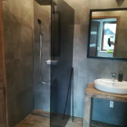 Nowoczesna łazienka z kabiną prysznicową typu walk-in, umywalką nablatową na drewnianym blacie i dużym lustrem w czarnej ramie, płytki w odcieniach szarości.