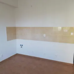 Wnętrze pomieszczenia z brązową podłogą z płytek i ścianami częściowo wyłożonymi beżowymi płytkami, widoczne puszki elektryczne i otwory na rury.