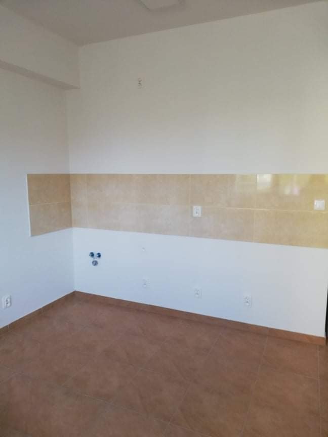 Wnętrze pomieszczenia z brązową podłogą z płytek i ścianami częściowo wyłożonymi beżowymi płytkami, widoczne puszki elektryczne i otwory na rury.