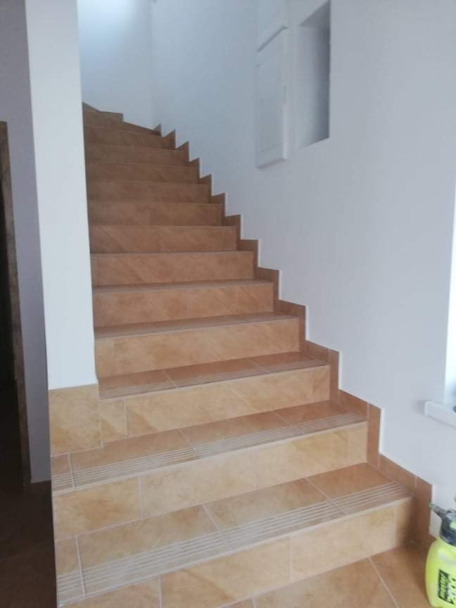 Wyłożone beżowymi płytkami schody wewnętrzne z antypoślizgowymi paskami na krawędziach stopni, widoczne z dołu, ściany pomalowane na biało, w rogu widoczny opryskiwacz.