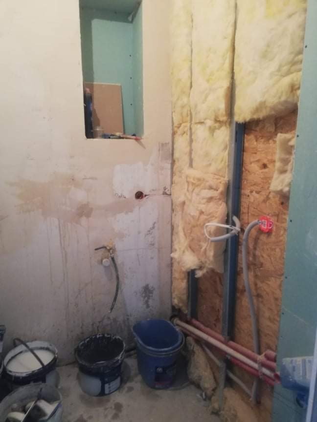 Wnętrze łazienki w trakcie remontu: ściana z częściowo ułożoną izolacją z wełny mineralnej, widoczne profile metalowe, instalacja wodna i elektryczna, ściana z nieukończonym tynkiem, wiadra...