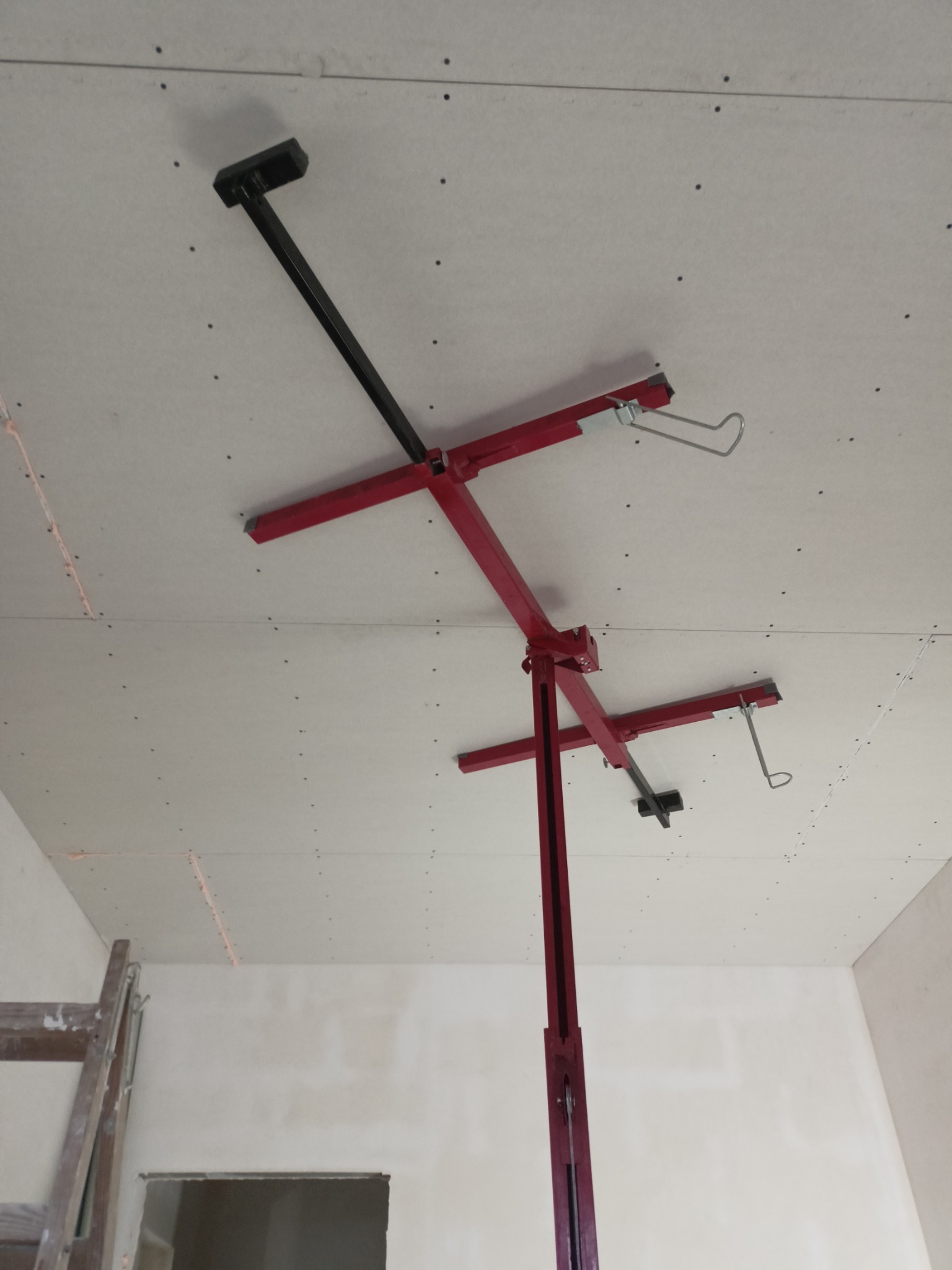 Czerwony podnośnik do płyt gipsowo-kartonowych podtrzymuje białą płytę sufitową, widoczne ślady po szpachlowaniu i wkrętach, drabina w rogu pomieszczenia.
