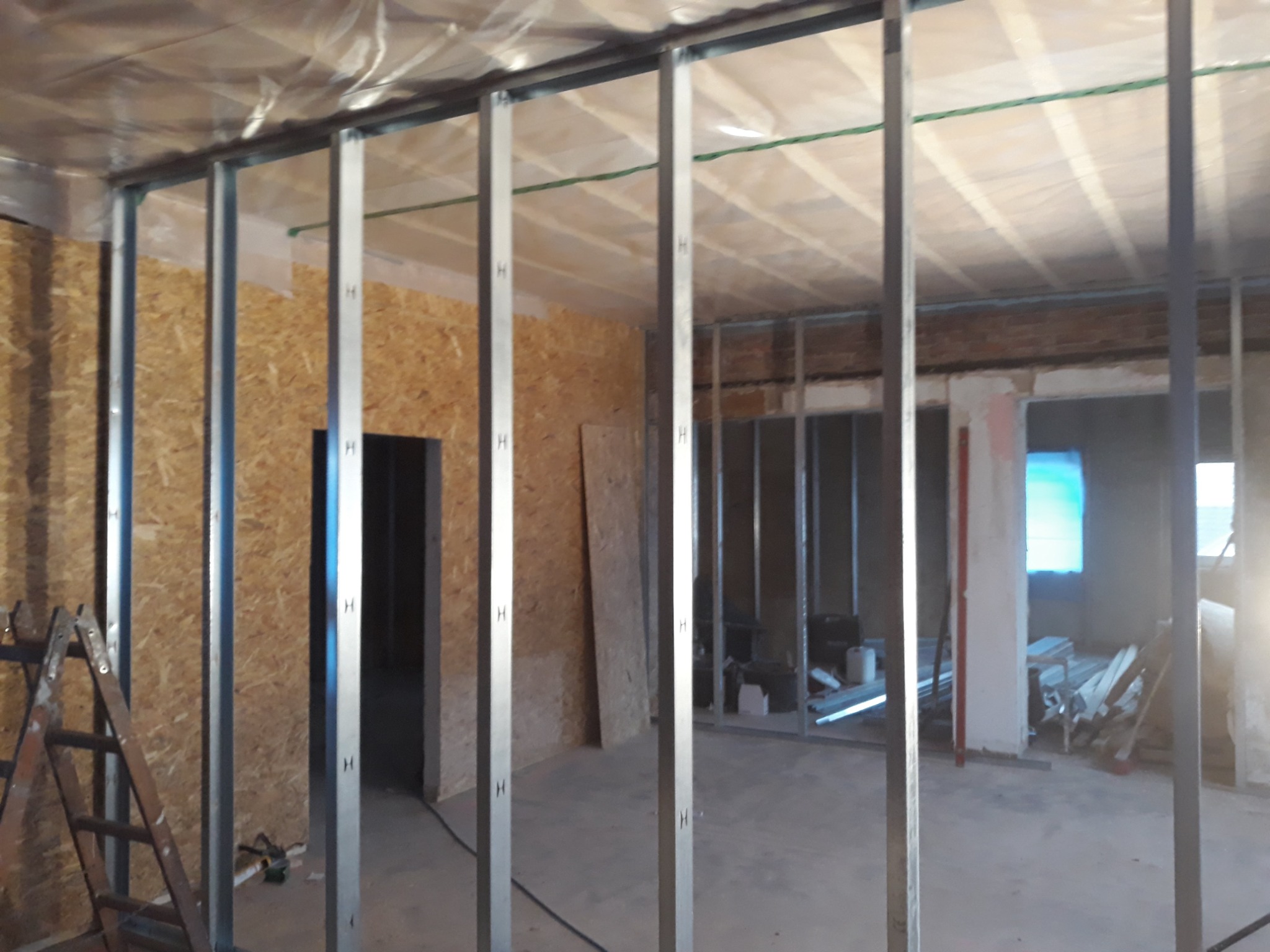 Wnętrze w trakcie remontu, widoczne metalowe profile konstrukcyjne ścian działowych, płyty OSB na ścianach, drabina malarska i folia zabezpieczająca sufit.