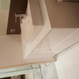 Biała, nowoczesna umywalka z szafką, zamontowana w łazience z błyszczącymi, białymi płytkami podłogowymi i podświetleniem LED.