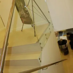 Szklana balustrada schodowa z metalową poręczą, mocowana punktowo do stopni, widok z góry, z odbiciem obrazu w szkle.