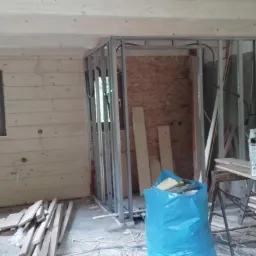 Wnętrze w trakcie remontu: drewniane ściany z oknem, metalowa konstrukcja ścianki działowej, drabina oparta o ścianę, narzędzia i materiały budowlane.