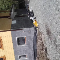 Dom w trakcie ocieplania: widoczna drewniana elewacja, fragment ściany z szarą izolacją, okna z drewnianymi ramami i folia kubełkowa przy fundamencie.