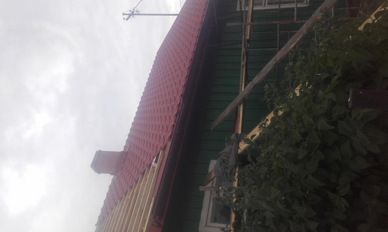 Częściowy widok domu z nową, czerwoną blachodachówką na dachu w trakcie montażu, widoczne elementy konstrukcyjne dachu, komin i fragment elewacji z oknem, pochmurne niebo w tle.