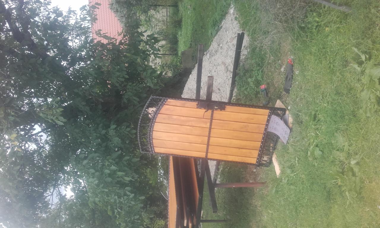 Element ogrodzenia z drewnianymi panelami i ozdobnymi, kutymi elementami, leżący na trawie obok ścieżki z płyt betonowych.