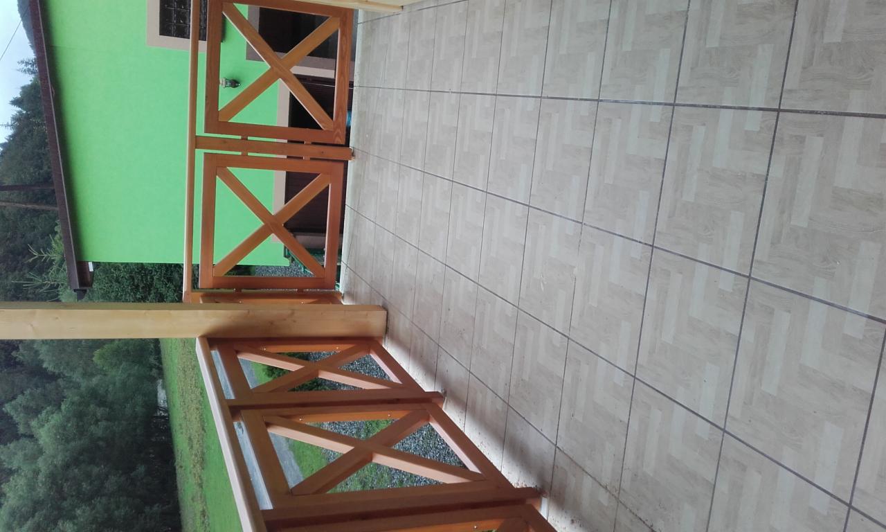 Wyłożony płytkami taras z drewnianą balustradą w kształcie krzyży, widok na zielony budynek i drzewa w tle.