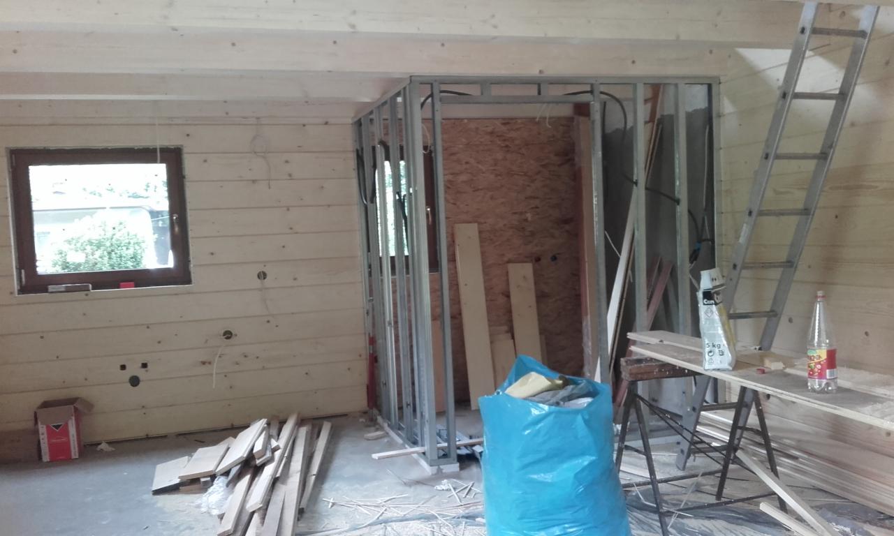 Wnętrze w trakcie remontu: drewniane ściany z oknem, metalowa konstrukcja ścianki działowej, drabina oparta o ścianę, narzędzia i materiały budowlane.
