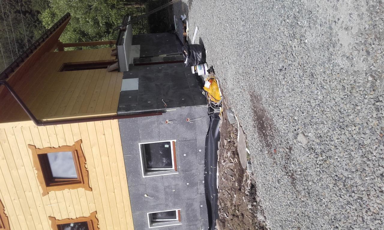 Dom w trakcie ocieplania: widoczna drewniana elewacja, fragment ściany z szarą izolacją, okna z drewnianymi ramami i folia kubełkowa przy fundamencie.