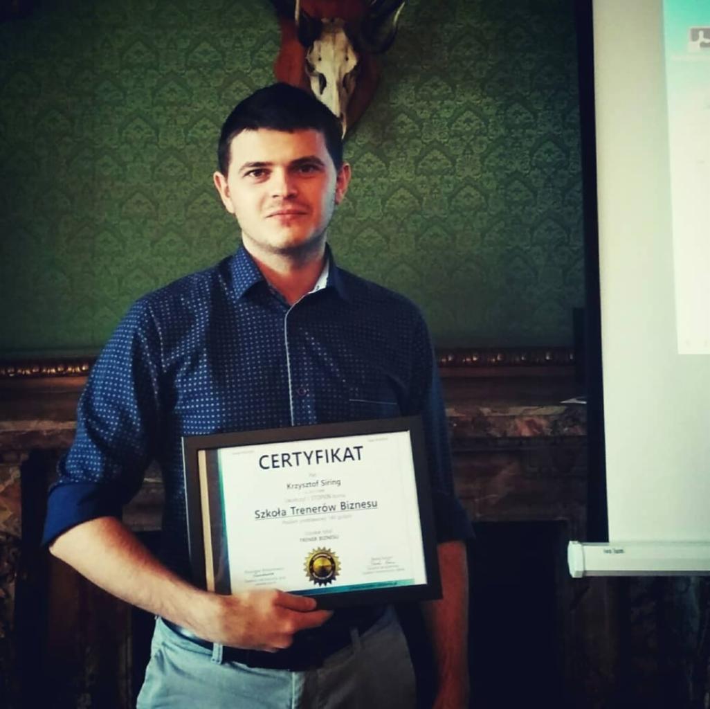 Mężczyzna trzyma w rękach oprawiony certyfikat ukończenia Szkoły Trenerów Biznesu, na ścianie zielona tapeta w ornamenty i trofeum myśliwskie.