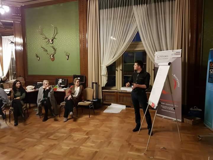 Prelegent prowadzi szkolenie w eleganckiej sali z dekoracjami myśliwskimi, flipchart z logo firmy i siedząca publiczność.