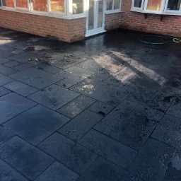 Świeżo ułożona, mokra, ciemnoszara kostka brukowa na tarasie przylegającym do domu z cegły i białymi oknami, widoczny zielony wąż ogrodowy.