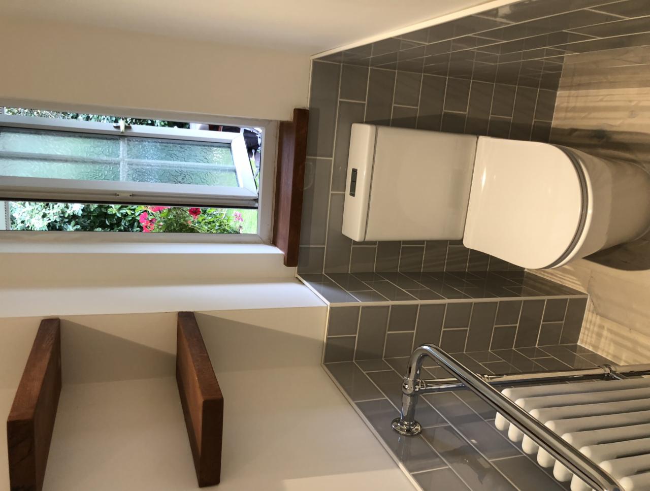 Nowoczesna toaleta z widokiem na ogród przez otwarte okno, szare płytki metra i drewniane półki.