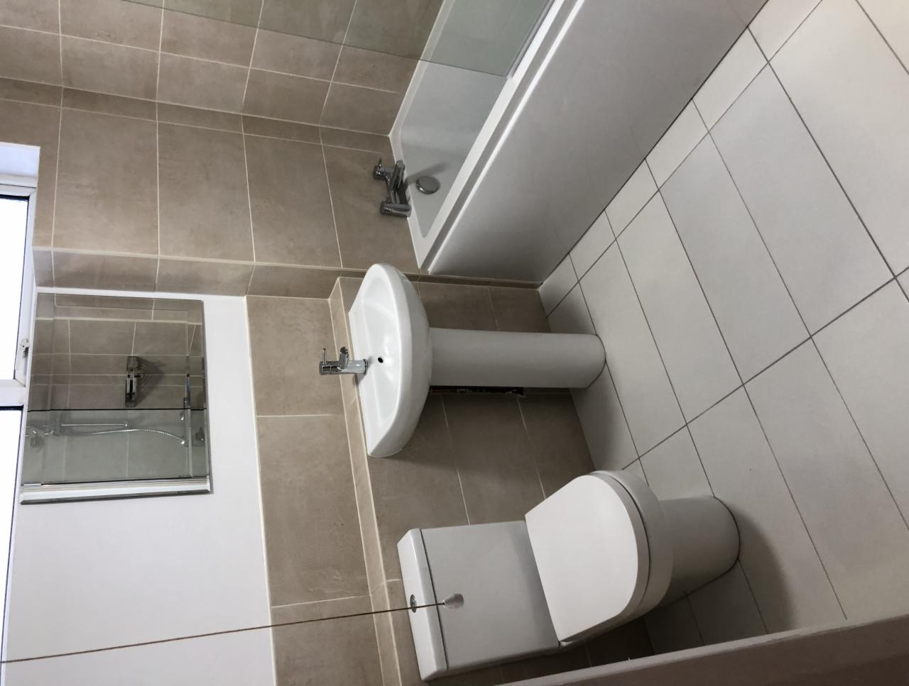 Jasna łazienka po remoncie z beżowymi płytkami, białą umywalką na postumencie, toaletą i wanną z parawanem.