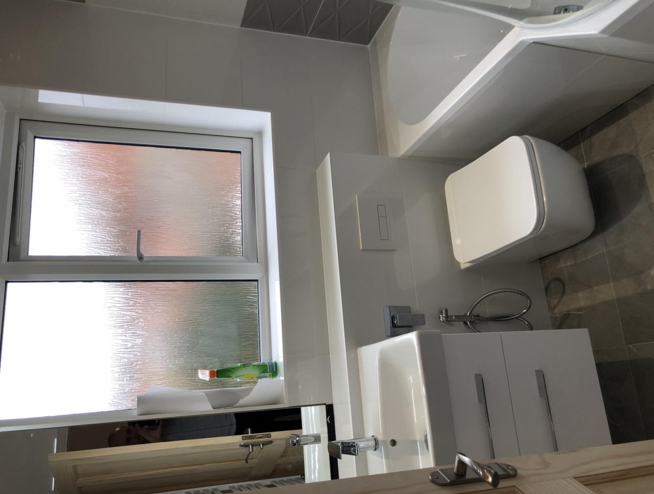 Nowoczesna łazienka z białą armaturą, umywalką z szafką, toaletą podwieszaną i wanną narożną, wykończona szarymi płytkami, z oknem z szybą ornamentową.