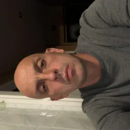 Portret mężczyzny w szarym swetrze, z bliska, z widoczną białą, kafelkową powierzchnią w tle.